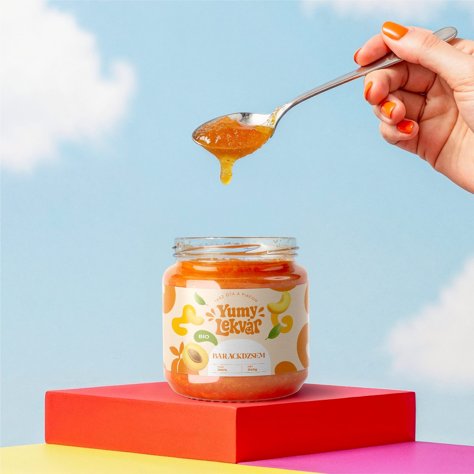 Spoon lifting apricot jam from a Yumy Lekvár jar on a red block under a blue sky: product shot by byannas
